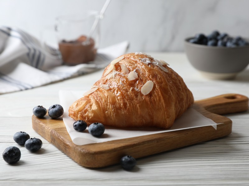 Ein gefülltes Mandelcroissant mit Mandeln und Puderzucker verziert auf einem Brett, rundherum Blaubeeren.