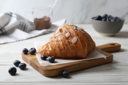 Ein gefülltes Mandelcroissant mit Mandeln und Puderzucker verziert auf einem Brett, rundherum Blaubeeren.