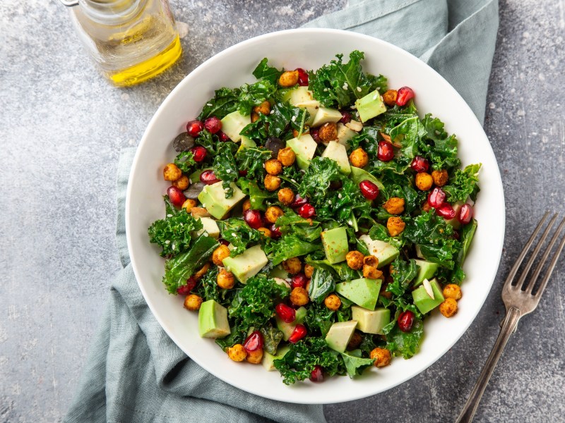 Eine Schüssel Grünkohlsalat mit Avocado und Kichererbsen sowie Granatapfelkernen in der Draufsicht, daneben eine Flasche Öl.