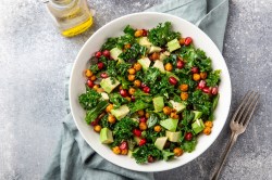 Eine Schüssel Grünkohlsalat mit Avocado und Kichererbsen sowie Granatapfelkernen in der Draufsicht, daneben eine Flasche Öl.