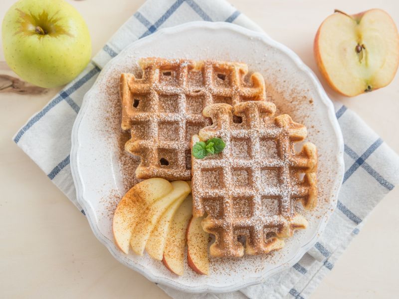 Haferflocken-Apfelmus-Waffeln angerichtet auf einem Teller mit Apfelspalten und Puderzucker. Der Teller steht auf einem Küchenhandtuch. Zwei Äpfel liegen als Dekoration neben dem Teller.