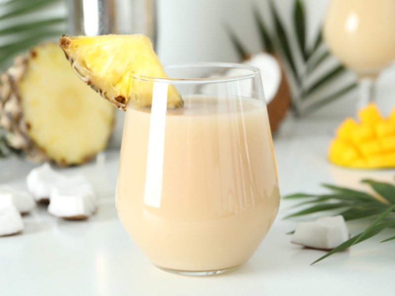 Ein Glas heiße Piña Colada mit einem Stück frischer Ananas als Deko vor Palmenblättern, frischer Ananas und Mango im Hintergrund.