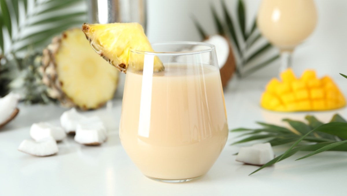 Ein Glas heiße Piña Colada mit einem Stück frischer Ananas als Deko vor Palmenblättern, frischer Ananas und Mango im Hintergrund.