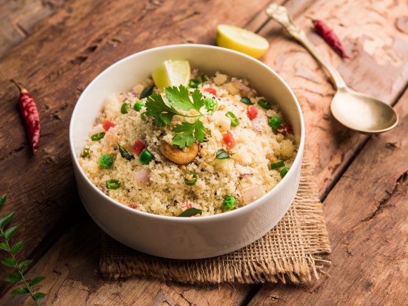 Eine Schale indisches Upma, Hartweizengrieß, mit etwas Gemüse und Kräutern, daneben ein Löffel.