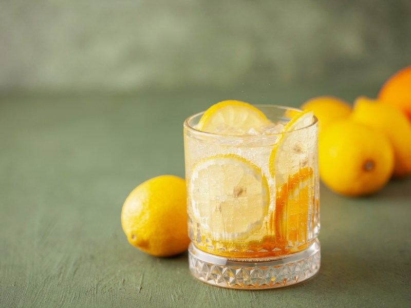 Ein Glas Limoncello Tonic mit Eiswüfeln und Zitronenscheiben, dahinter frische Zitronen.