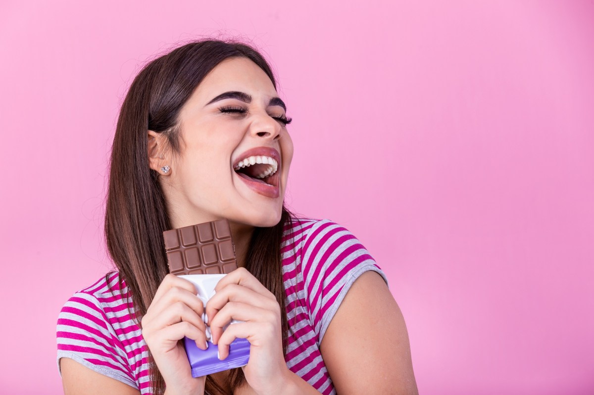 Macht Schokolade glücklich? Eine fröhlich lachende dunkelhaarige junge Frau im gestreiften T-Shirt hält eine Tafel Schohkolade in der Hand, rosafarbener Hintergrund.