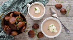 Zwei kleine Suppenterrinen mit Maronen-Cremesuppe, die mit Thymian und einem Klecks Soja-Kochcreme garniert sind. daneben steht eine Schale mit Marone und es liegen einige drumherum.