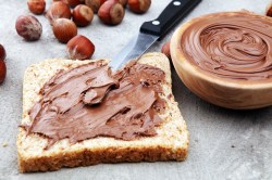 Selbst gemachte Nutella auf einer Toastbrotscheibe mit einem Messer, daneben eine Schale mit der Nutella und Haselnüsse.