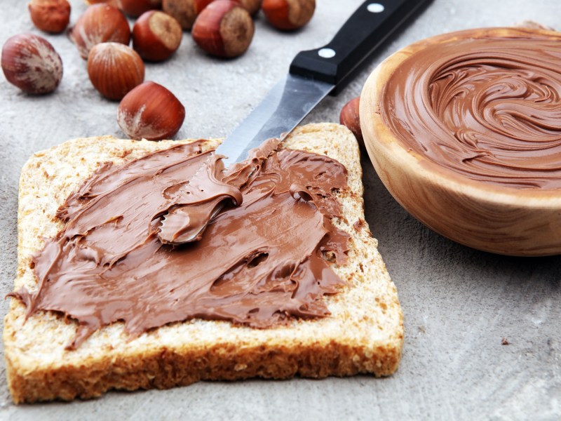 Selbst gemachte Nutella auf einer Toastbrotscheibe mit einem Messer, daneben eine Schale mit der Nutella und Haselnüsse.