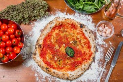 Pizza Marinara mit Tomatensoße, Knoblauch und einem Blatt Basilikum in der Draufsicht, daneben eine Schale Tomaten, getrockneter Oregano, Basilikum und Besteck,