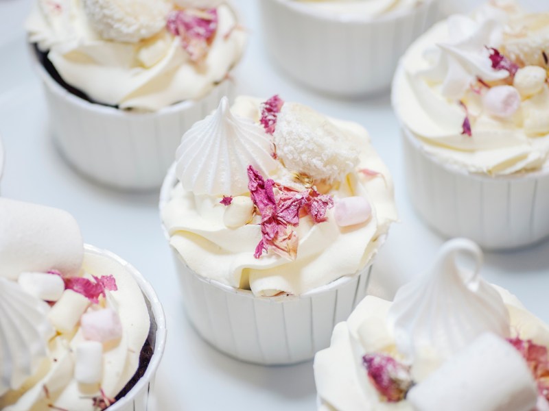 Prosecco-Cupcakes auf einem weißen Untergrund, dekoriert mit Rosenblättern, Marshmallows und Baiser.