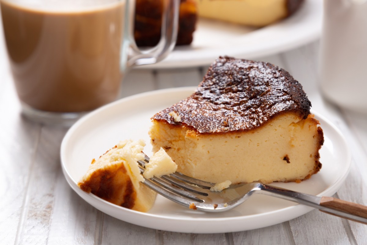 Ein Stück Quarkkuchen ohne Boden auf einem Teller, darauf auch eine Kuchengabel, im Hintergrund unscharf ein Glas Kaffee.