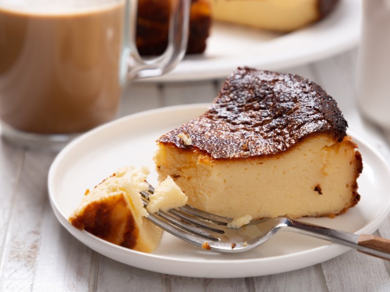 Ein Stück Quarkkuchen ohne Boden auf einem Teller, darauf auch eine Kuchengabel, im Hintergrund unscharf ein Glas Kaffee.