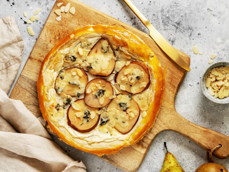Schnelle Blätterteig-Birnentarte mit Mandeln auf einem Brett, daneben ein goldenes Messer und ein paar Zutaten.