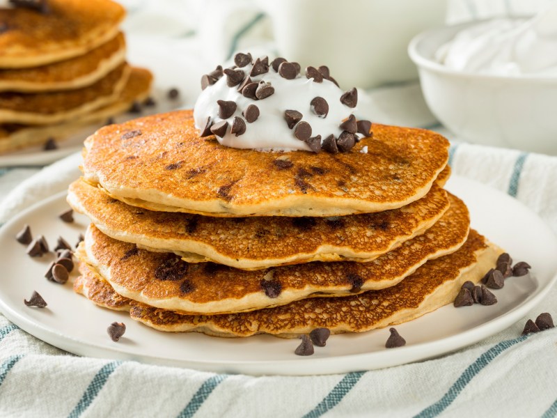 Ein Stapel Schoko-Bananen-Pancakes mit Schokodrops und Sahne serviert, auf einem Teller, auf einem gestreiften Tischtuch.