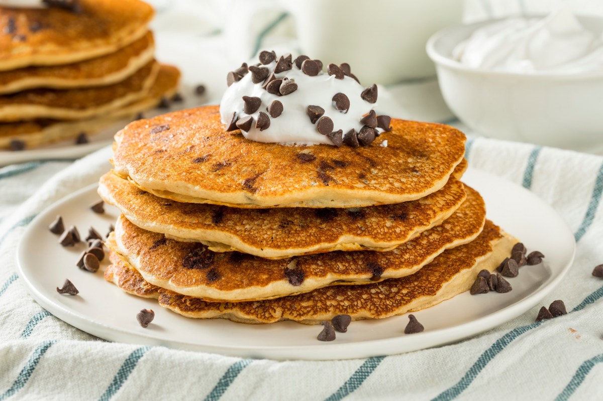 Ein Stapel Schoko-Bananen-Pancakes mit Schokodrops und Sahne serviert, auf einem Teller, auf einem gestreiften Tischtuch.