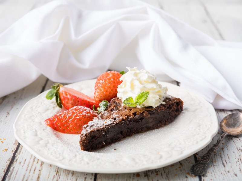 Ein Stück schwedischer Schokoladenkuchen Kladdkaka auf einem Teller, garniert mit Erdbeeren und Sahne, daneben ein Löffel.