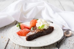 Ein Stück schwedischer Schokoladenkuchen Kladdkaka auf einem Teller, garniert mit Erdbeeren und Sahne, daneben ein Löffel.