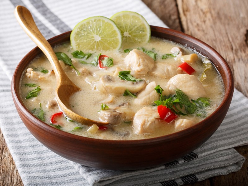 Tom Kha Gai Suppe in einer braunen Schale mit einem Holzlöffel. Sie ist mit zwei Limettenscheiben dekoriert.