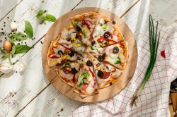 Eine Tortilla-Pizza mit Oliven, Pilzen, Mais und Paprika sowie Käse in der Draufsicht, daneben frische Zutaten.