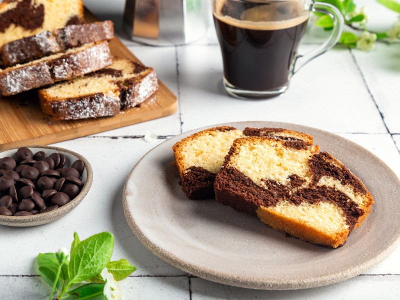 2 Stück Marmorkuchen auf einem Teller, daneben eine Schale Schokodrops, der angeschnittene Kastenkuchen auf einem Brett und eine Tasse Kaffee.