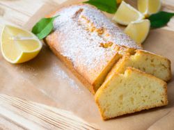 Aufgeschnittener veganer Zitronenkuchen, serviert auf einem Brett. Eine aufgeschnittene Zitrone und Minzblätter liegen als Deko neben dem Kuchen.