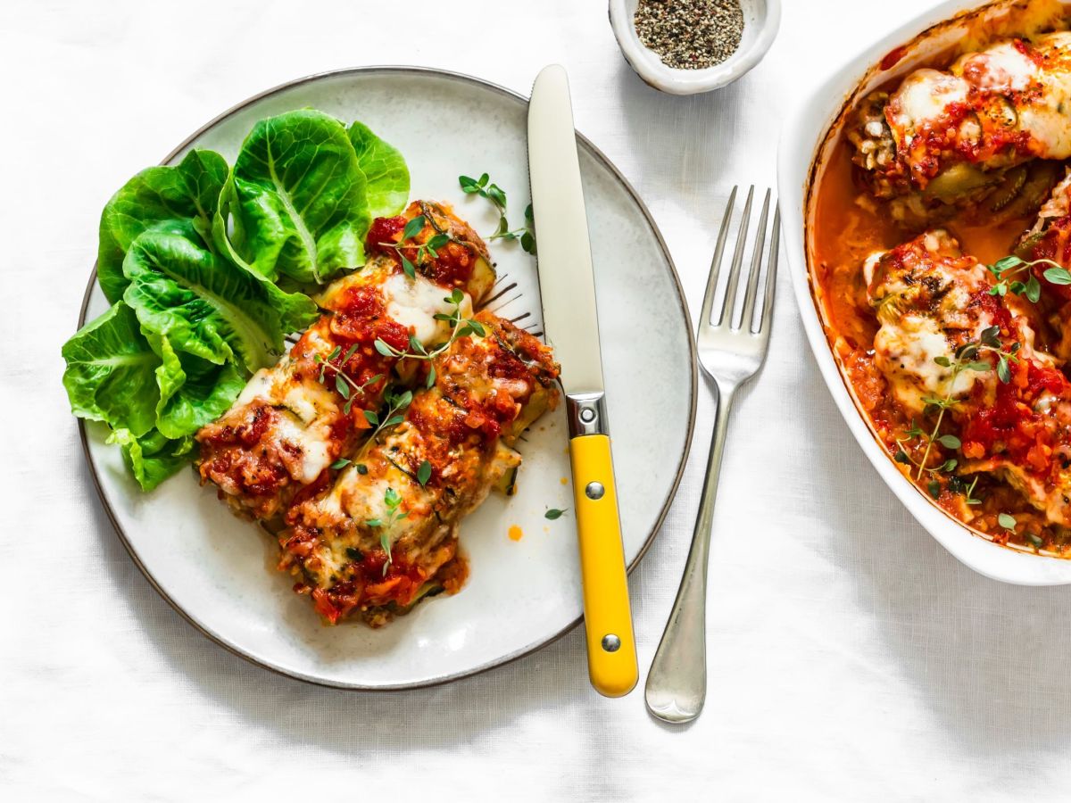 Zucchini-Enchiladas, angerichtet auf einem Teller mit Salat, daneben liegt Besteck. Daneben steht eine Form mit Zucchini-Enchiladas .