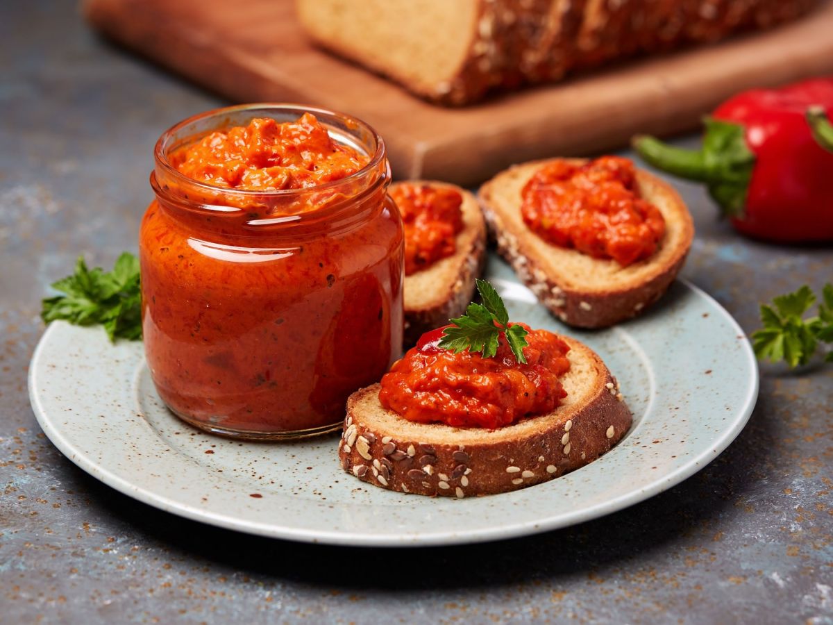 Ein Glas mit Ajvar-Feta-Dip steht auf einem Teller, daneben liegen mehrere Brotscheiben mit dem Dip. Im Hintergrund liegen Brot und Paprika als Dekoration.