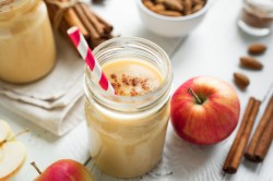 Ein Glas Apfel-Bananen-Smoothie mit Zimt bestreut, darin ein Strohhalm, drumherum Zimtstangen und Äpfel.