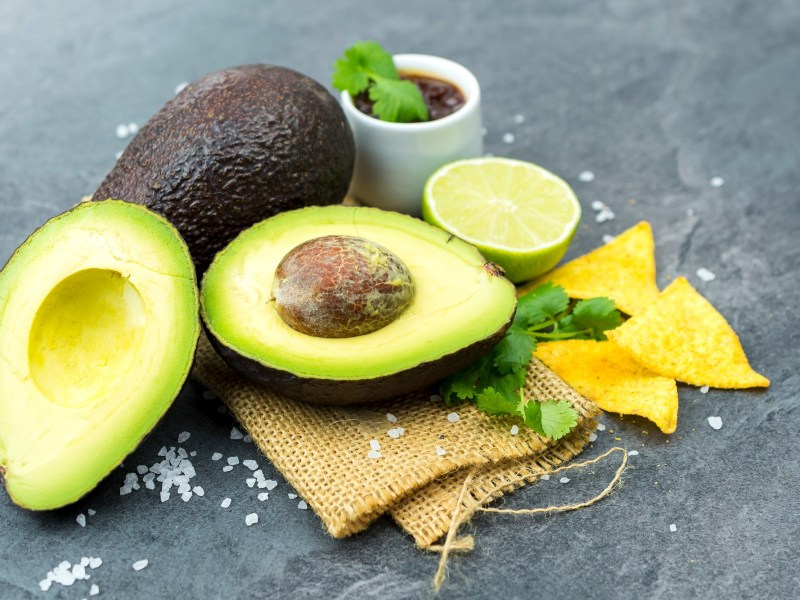 Avocado schneller reifen lassen: Eine ganze und eine halbierte Avocado, daneben Nachos, eine halbe Limette und ein Schälchen mit Dip.