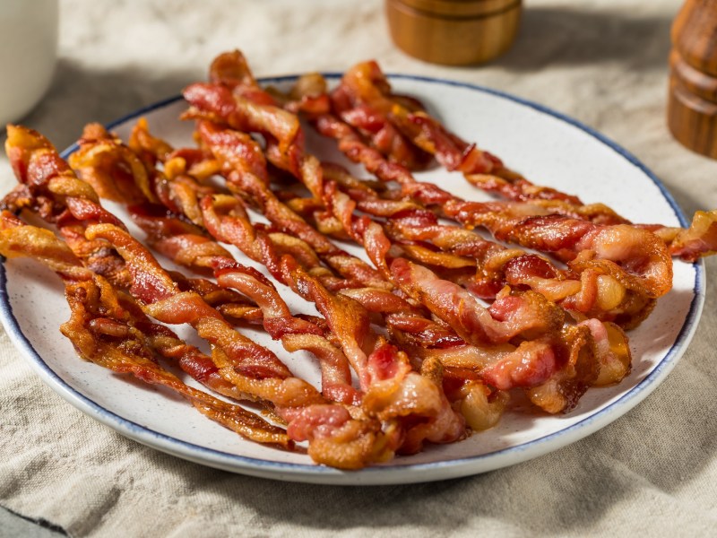 Bacon-Zöpfe auf einem weißen Teller, der auf einer beigen Tischdecke steht.
