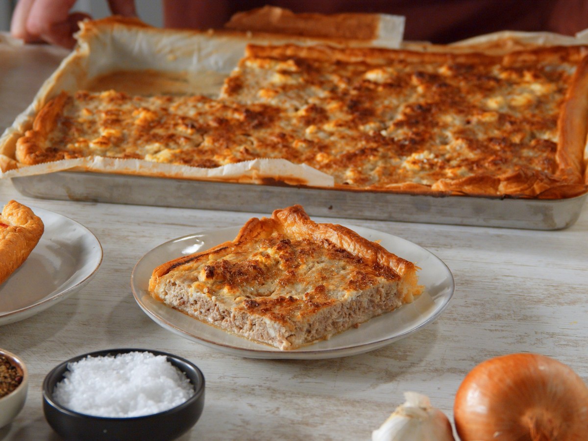 Ein Stück Blätterteig-Quiche mit Hackfleisch, Sahne und Ei auf einem Teller, dahinter das Backblech mit der Quiche.