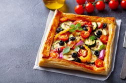 Blätterteigpizza mit mediterranem Gemüse auf einem weißen Backpapier Daneben liegen eine Rispe Kirschtomaten und ein Messer. Im Hintergrund steht ach eine Schale Olivenöl.