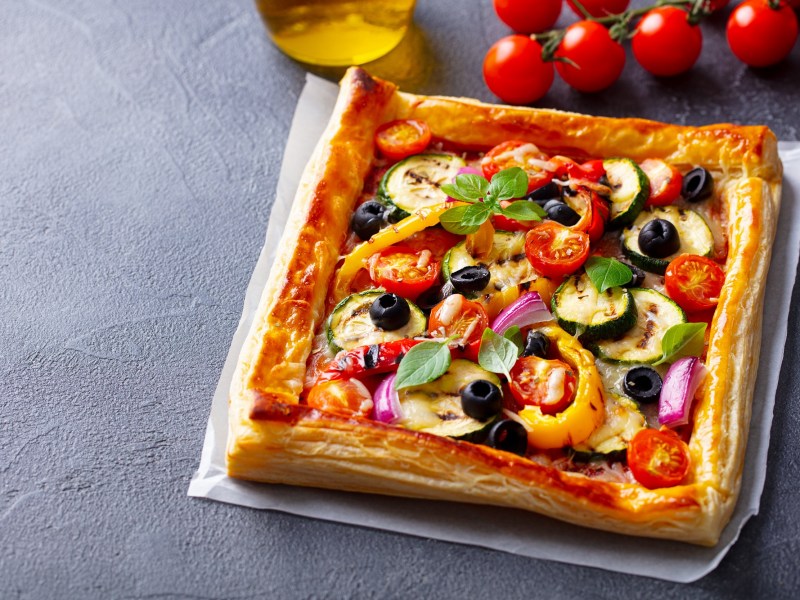 Blätterteigpizza mit mediterranem Gemüse auf einem weißen Backpapier Daneben liegen eine Rispe Kirschtomaten und ein Messer. Im Hintergrund steht ach eine Schale Olivenöl.