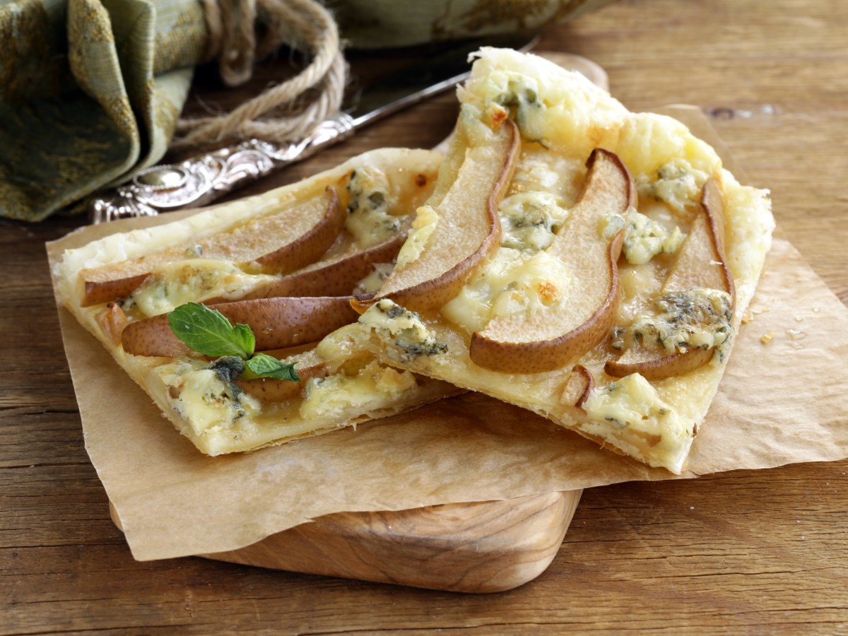 Zwei Stücke Blätterteig-Tarte mit Birne und Blauschimmelkäse auf Backpapier und einem Holzbrett.