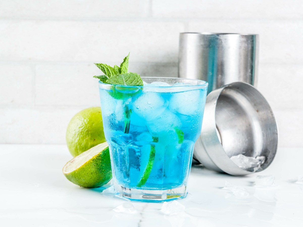Ein Glas Blue Lagoon mit Eiswürfeln, Limettenscheiben und Minze garniert, dahinter ein Cocktailshaker und halbierte Limetten.
