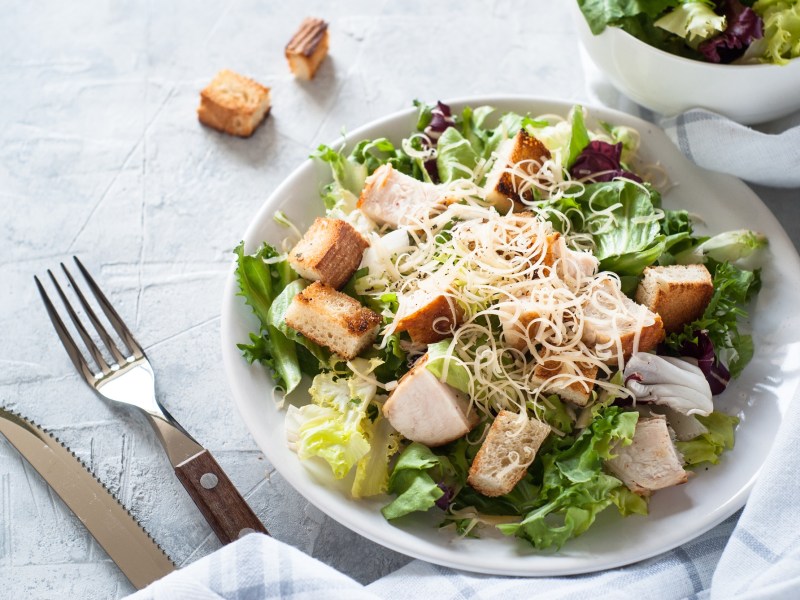 Teller mit Caesar Salat und Besteck auf hellem Untergrund