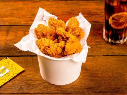 Crispy Chicken Wings, serviert in einem Eimer. Im HIntergrund steht eine Cola und liegt ein Tütchen Senf.