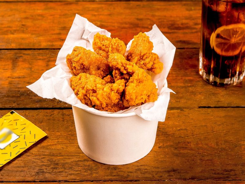 Crispy Chicken Wings, serviert in einem Eimer. Im HIntergrund steht eine Cola und liegt ein Tütchen Senf.