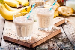 2 Gläser Erdnussbutter-Bananen-Smoothie mit Strohhalm und Haferflocken auf einem Brett, drumherum Bananen und Haferflocken.