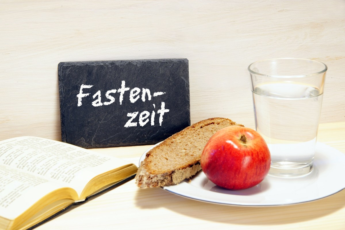 Fasten nach Karneval: ein aufgeschlagenes Buch, eine kleine Tafel mit der Aufschrift "Fastenzeit" sowie ein Teller mit einem Apfel und einer Scheibe Brot, daneben ein Glas Wasser.