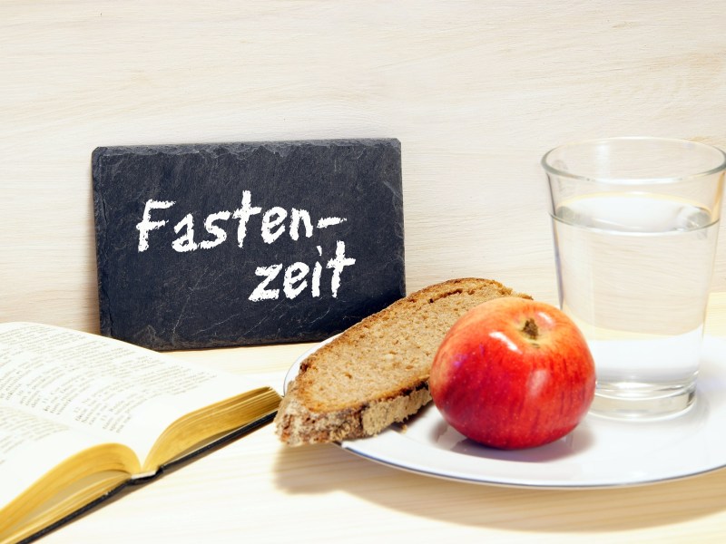 Fasten nach Karneval: ein aufgeschlagenes Buch, eine kleine Tafel mit der Aufschrift "Fastenzeit" sowie ein Teller mit einem Apfel und einer Scheibe Brot, daneben ein Glas Wasser.