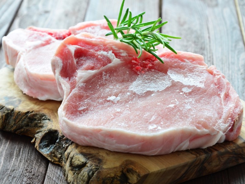 2 Schweine-Steaks auf einem Holzbrett, garniert mit einem Rosmarinzweig. Das Fleisch wurde eingefroren und aufgetaut.