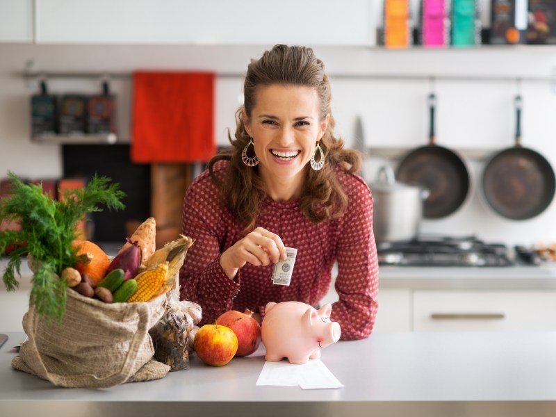 Geld sparen beim Kochen: eine lachende, lockige Frau steht in der Küche, steckt einen Geldschein in ein Sparschwein, neben ihr liegen Einkäufe.