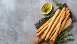 Gedrehte Grissini auf einem Holzbrett, daneben eine Schale mit Olivenöl uns Rosmarin