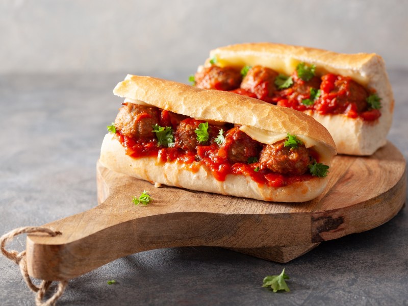 2 Italienische Meatball-Sandwiches mit Käse und Petersilie und Tomatensoße auf einem Brett.