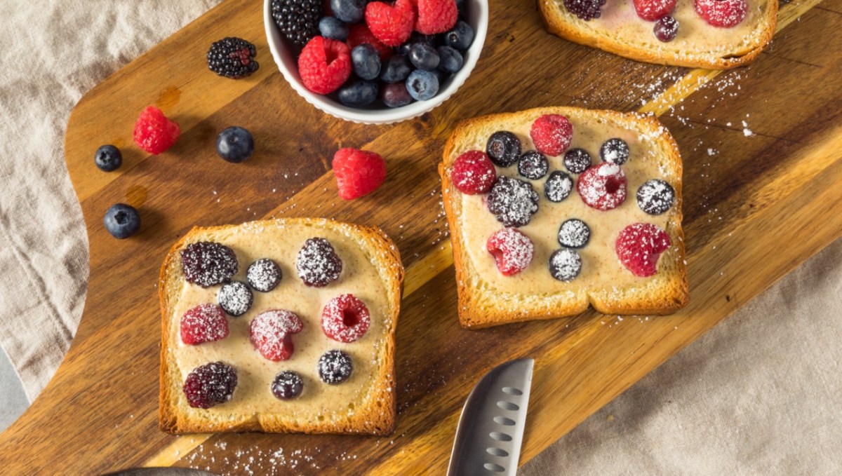 2 Scheiben Joghurt-Toast mit frischen Beeren auf einem Holzbrett. Daneben steht eine Schale mit Früchten und es liegt ein Messer am unteren Rand.