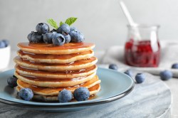 Ein Stapel Joghurt-Zitronen-Pancakes mit Blaubeeren und Minze garniert auf einem Teller, him Hintergrund ein Marmeladenglas.