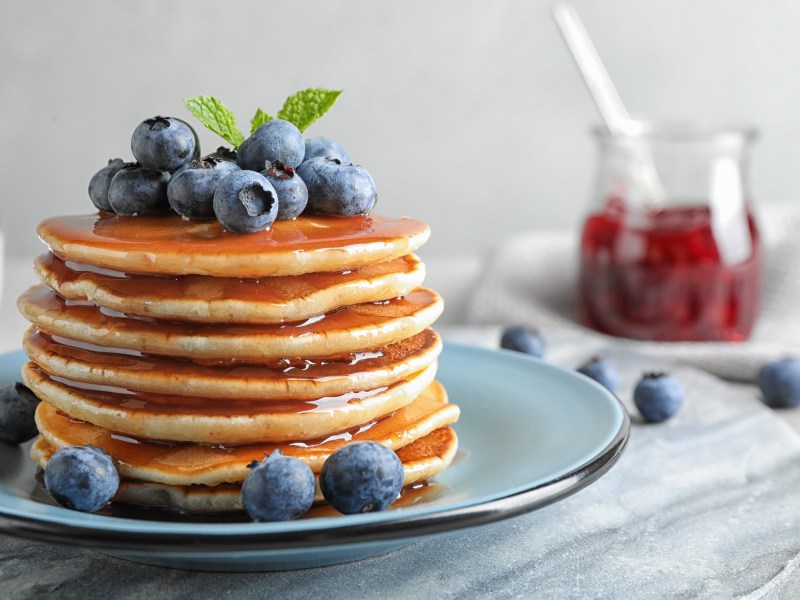 Ein Stapel Joghurt-Zitronen-Pancakes mit Blaubeeren und Minze garniert auf einem Teller, him Hintergrund ein Marmeladenglas.