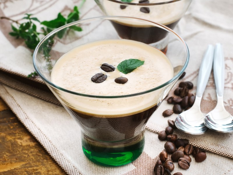 Kaffee-Zitronen-Shake in einem klassischen Glas, garniert mit Minze und Kaffeebohnen. Daneben zwei Löffel und noch mehr Kaffeebohnen.